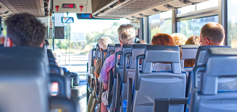 Passengers seated on a sunny bus ride. View from the back shows diverse individuals looking forward. Bright light filters in through large windows.