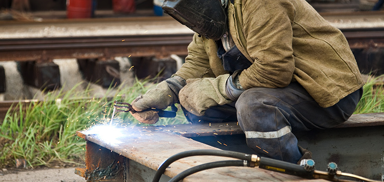 A welder in protective gear kneels on a metal structure, creating bright sparks as they work. Railroad tracks and green grass are in the background.