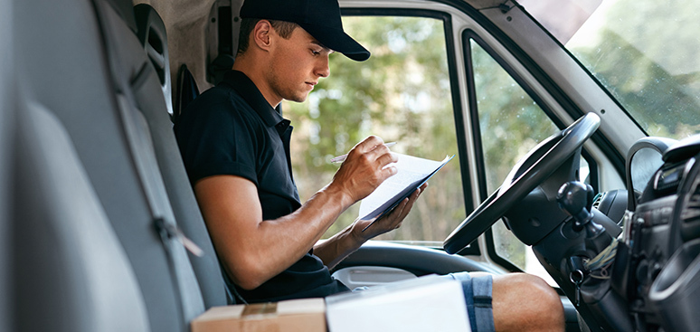A delivery driver in a cap focuses intently on paperwork inside a van. He sits next to packages, conveying concentration and diligence.