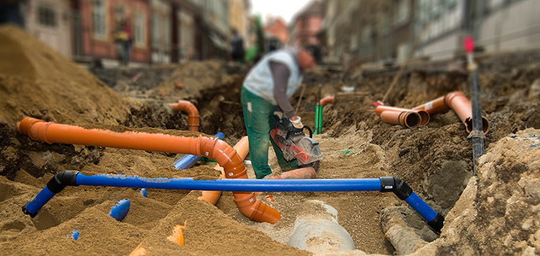 A construction worker in a trench adjusts orange and blue pipes amidst sand and dirt, with blurred buildings in the background, conveying a sense of urban infrastructure repair.
